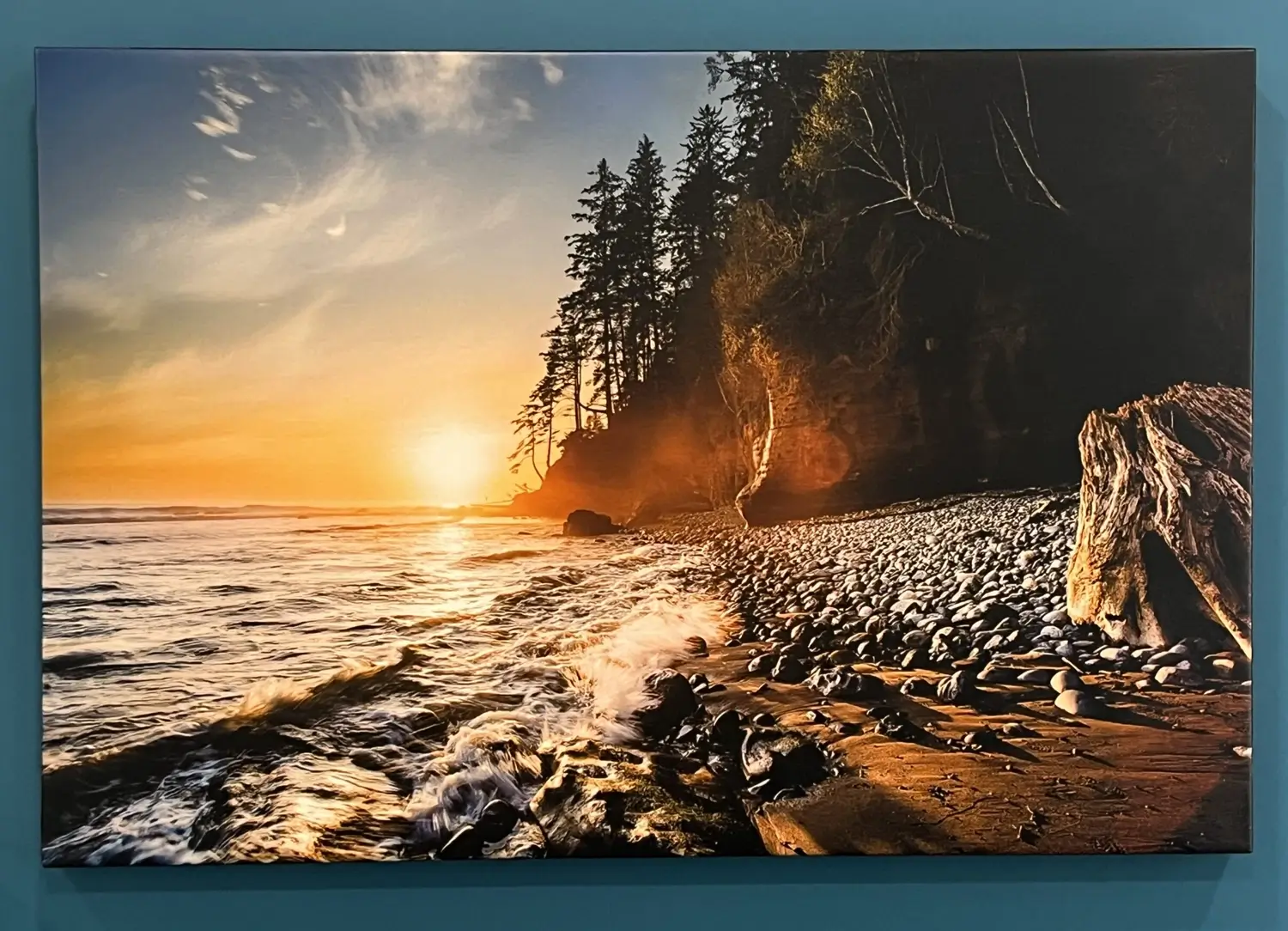 A large wall art print of a rocky beach at sunset, with waves crashing onto the shore and tall evergreen trees silhouetted against a golden sky.