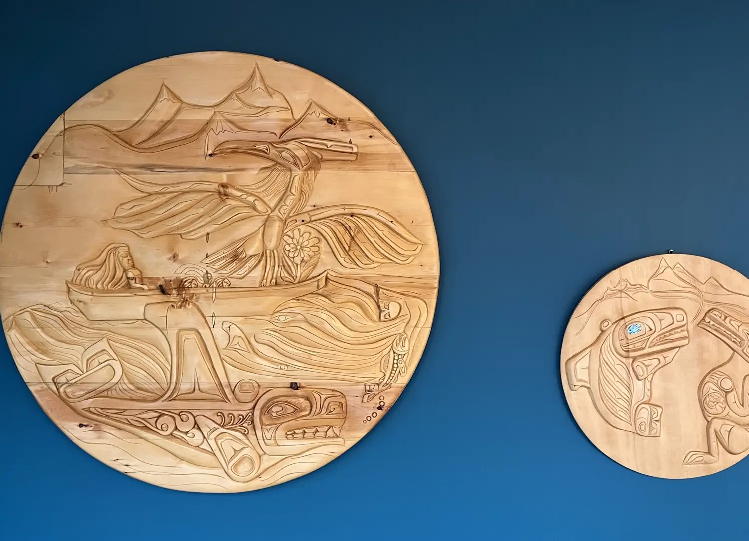 Two round wooden carvings featuring Indigenous Northwest Coast–style artwork hung on a deep blue wall; the larger carving shows a person in a canoe, a whale tail, and flowing water, while the smaller carving depicts traditional animal figures.