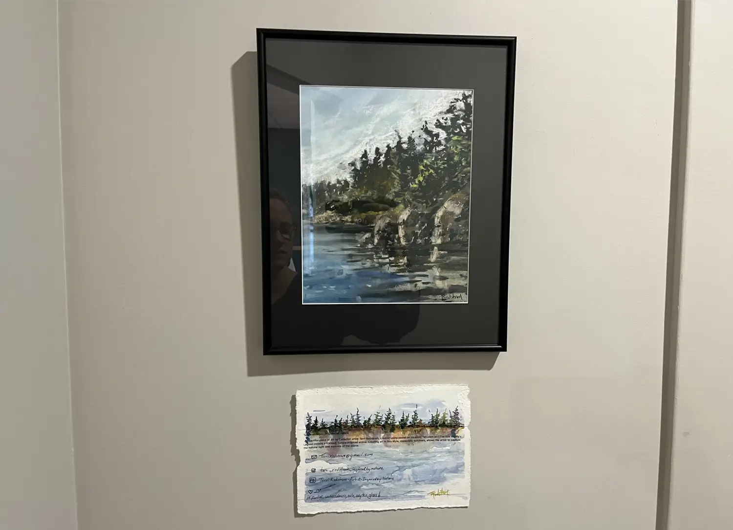 Framed coastal landscape painting depicting rocky shoreline, evergreen trees, and reflective water, displayed above a smaller watercolor of a similar tree-lined shoreline on textured paper.