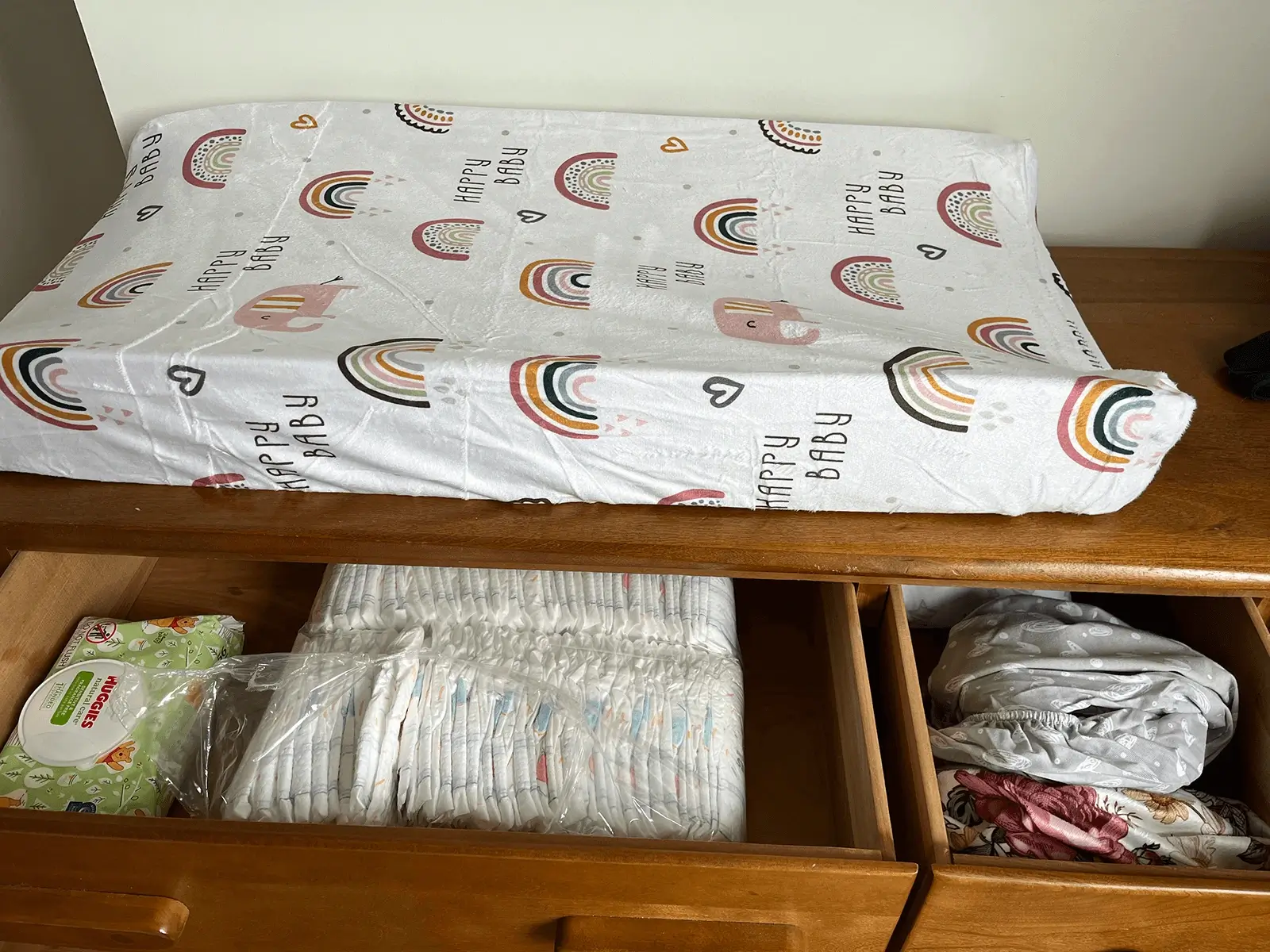 A baby changing station with a rainbow-patterned changing pad on a wooden dresser, with drawers below stocked with diapers, wipes, and folded baby linens.