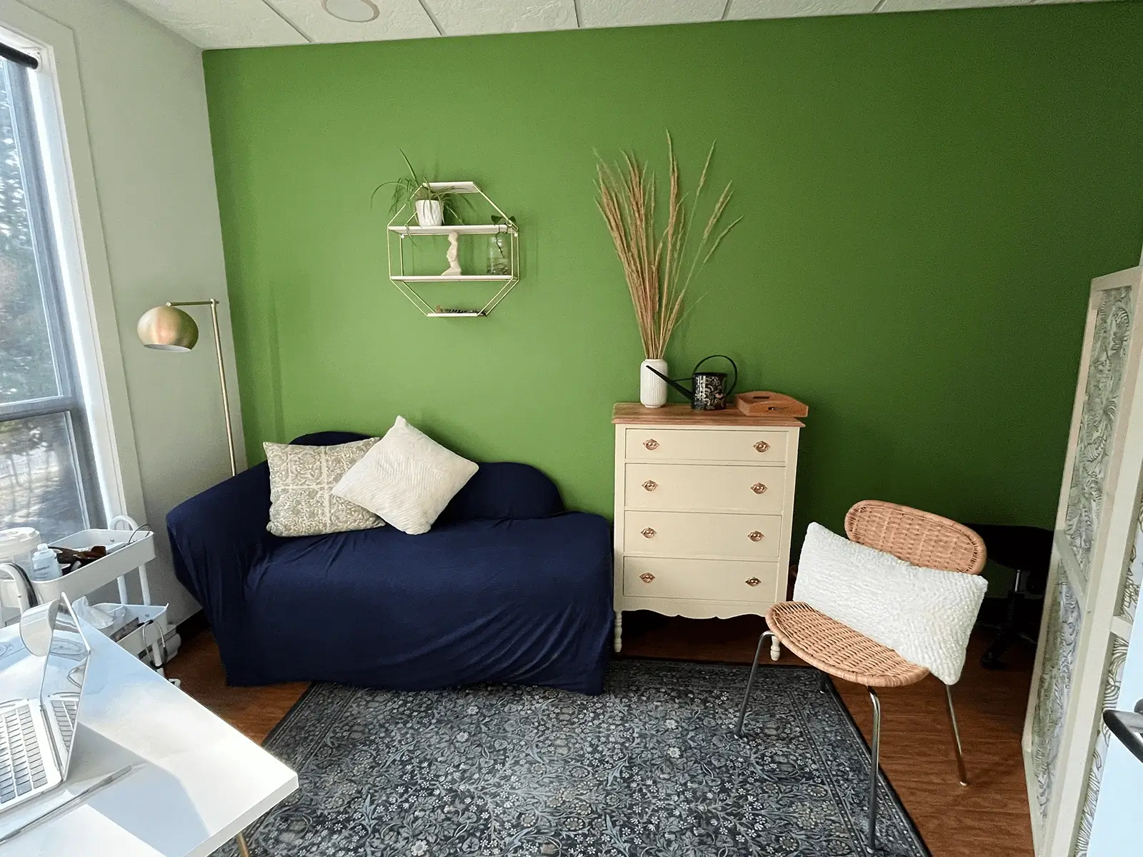 A cozy consultation room with a navy loveseat, patterned pillows, a cream dresser, a woven chair, and decor against a green accent wall.