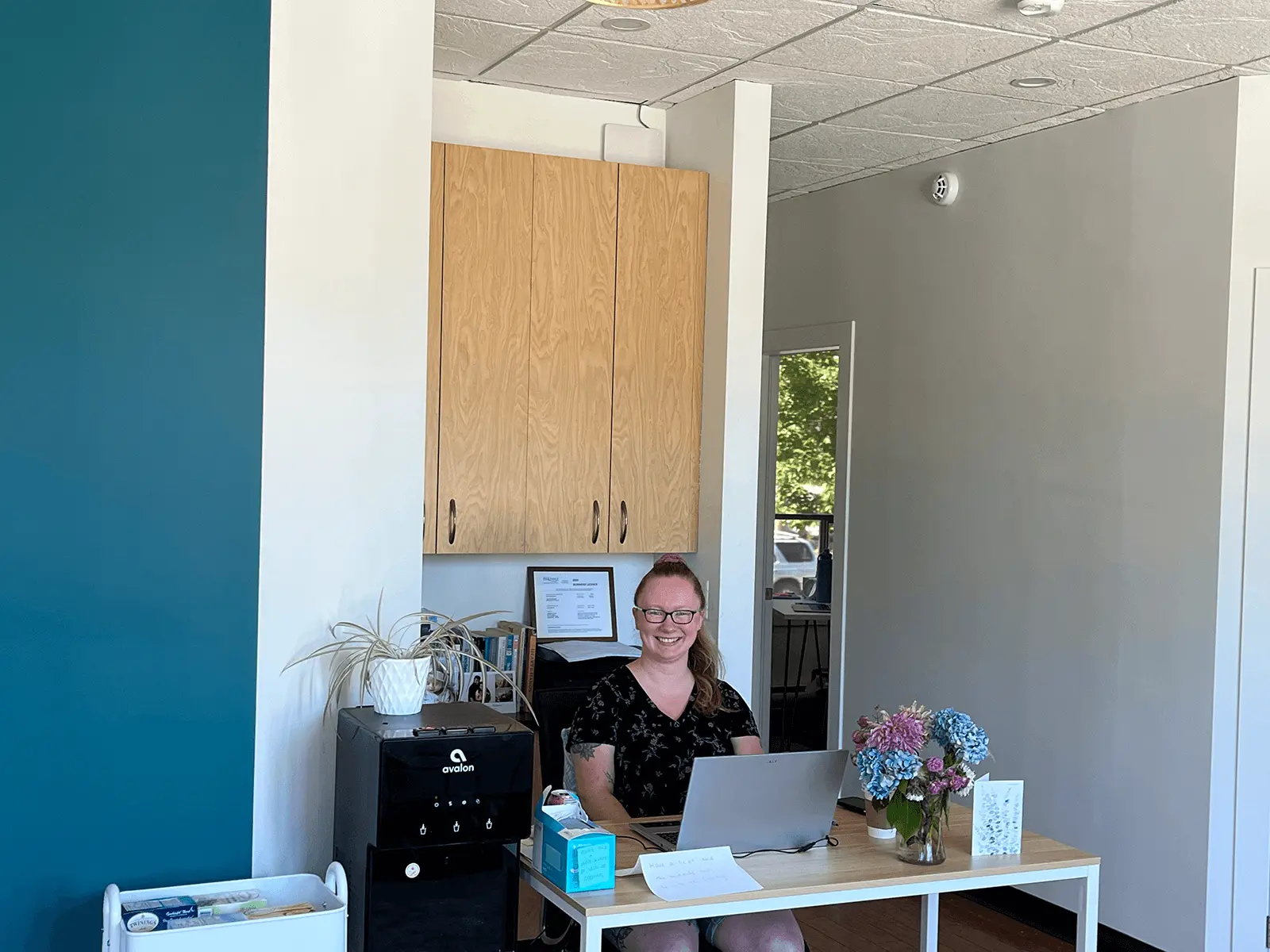 Reception area with a smiling staff member seated at a desk with a laptop, flowers, and paperwork, set against teal and gray walls and wooden cabinetry.
